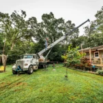 White crane truck performing tree removal in a residential backyard with green lawn, house, and garden features