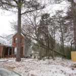 Fallen tree on residential property after winter storm, showing snow-covered yard, brick house exterior, and need for professional tree removal services